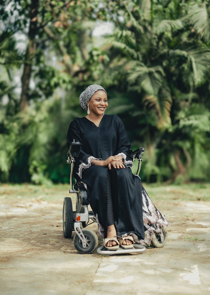 A woman sitting in a wheelchair, smiling outdoors with lush greenery around.